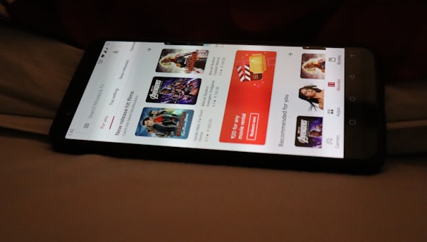 A smartphone displaying a streaming app interface, showing various movie and TV show thumbnails. The screen includes categories like 'For you' and 'New release hit films', with images from popular films. The background is a dimly lit setting with a fabric surface.