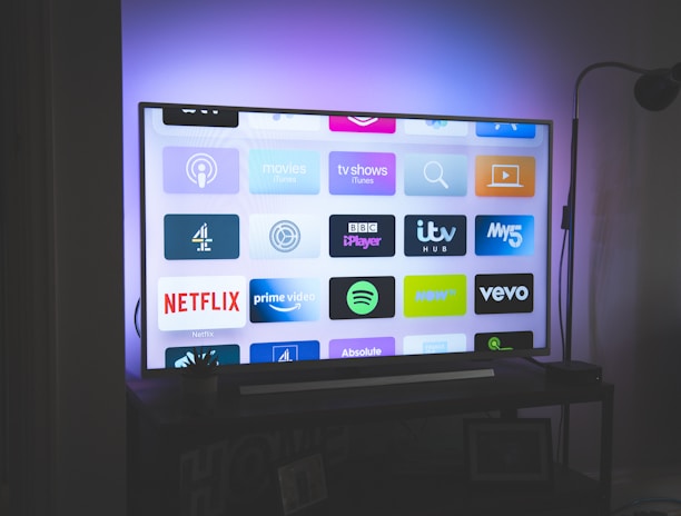 A flat-screen television is displayed in a dimly lit room, showcasing a variety of streaming service apps on its screen, such as Netflix, Prime Video, and Spotify. The TV is positioned on a black stand beside a tall floor lamp, casting a soft ambient light on the surrounding area.