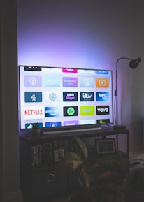 A flat-screen television is displaying a variety of streaming app icons, including Netflix, Prime Video, Spotify, and ITV Hub among others. The TV is placed on a media console with a small potted plant beside it. A tall lamp stands to the right, and a faint, ambient light casts a purple hue on the wall above the TV.