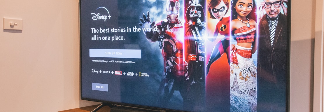 A television displaying a Disney+ promotional screen featuring various characters from popular franchises. Below the screen is a wooden cabinet with a few books and a small plant. The wall is light-colored, creating a cozy and inviting atmosphere.
