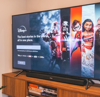 A television displaying a Disney+ promotional screen featuring various characters from popular franchises. Below the screen is a wooden cabinet with a few books and a small plant. The wall is light-colored, creating a cozy and inviting atmosphere.