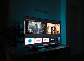 A television screen displaying a user interface with popular streaming service apps including Netflix, Sky, Apple TV, Prime Video, and others. The screen also shows images from movies or shows above the apps. The room is dimly lit with a potted plant visible on the side.