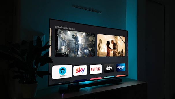 A television screen displaying a user interface with popular streaming service apps including Netflix, Sky, Apple TV, Prime Video, and others. The screen also shows images from movies or shows above the apps. The room is dimly lit with a potted plant visible on the side.