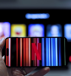A hand holding a smartphone displaying the Netflix logo on its screen with a background of colorful vertical lines. In the background, a television screen shows blurry icons or profiles with various colors.