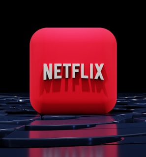 A red, cube-shaped icon with the word 'NETFLIX' prominently displayed in white, bold letters. The cube is set against a dark, glossy, circular patterned background, reflecting a sleek, futuristic design.