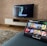 A modern living room features a flat-screen television mounted on a light-colored wooden stand with a patterned wall backdrop. Below the TV, a carpet lies on a wooden floor. In the foreground, a tablet displays a streaming platform interface with various show thumbnails, suggesting a relaxed entertainment setup.