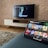 A modern living room features a flat-screen television mounted on a light-colored wooden stand with a patterned wall backdrop. Below the TV, a carpet lies on a wooden floor. In the foreground, a tablet displays a streaming platform interface with various show thumbnails, suggesting a relaxed entertainment setup.